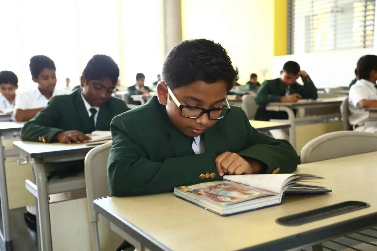 A student, inside the DPS Warangal school classroom, wearing a school uniform, and glasses, reading a book.