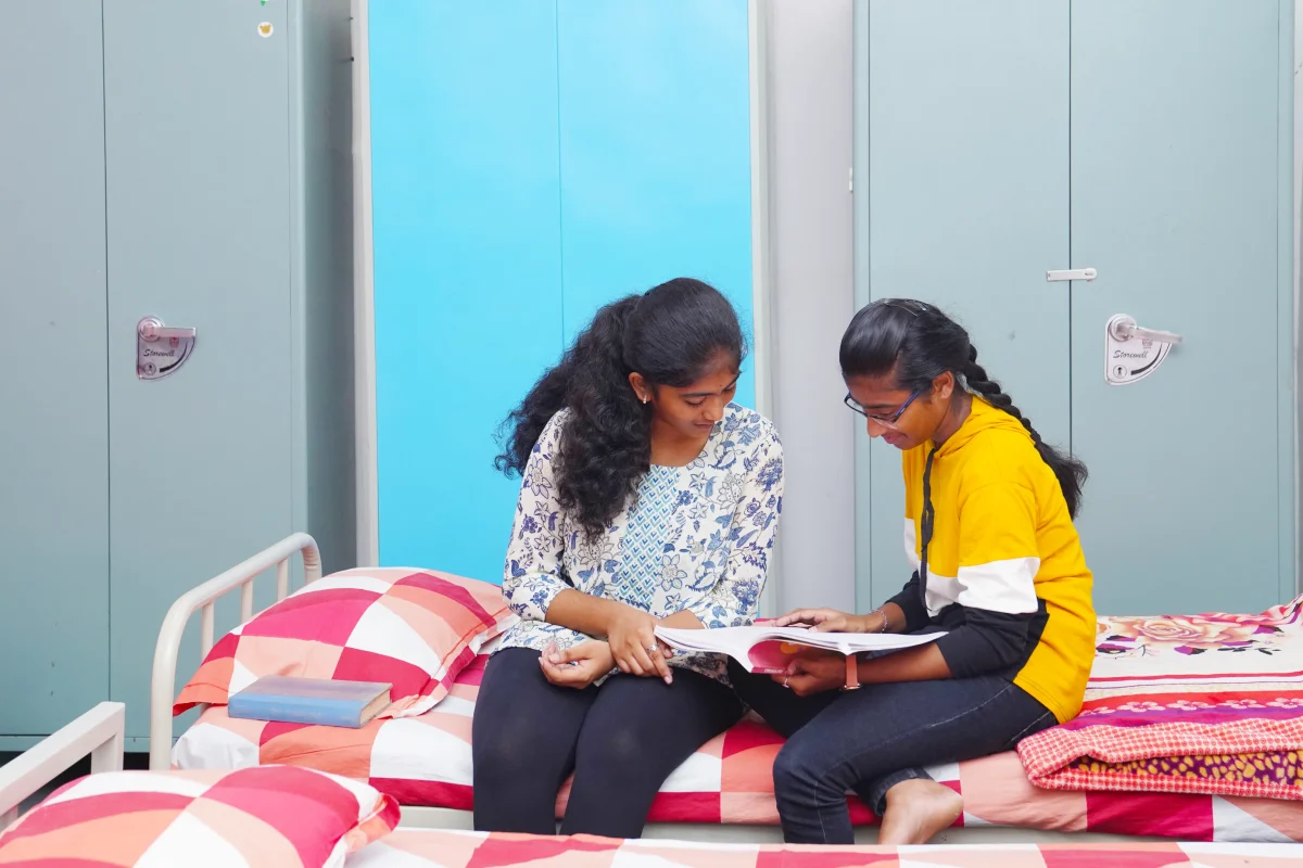 Two girls sitting in the hostel next to each other, helping in their studies.