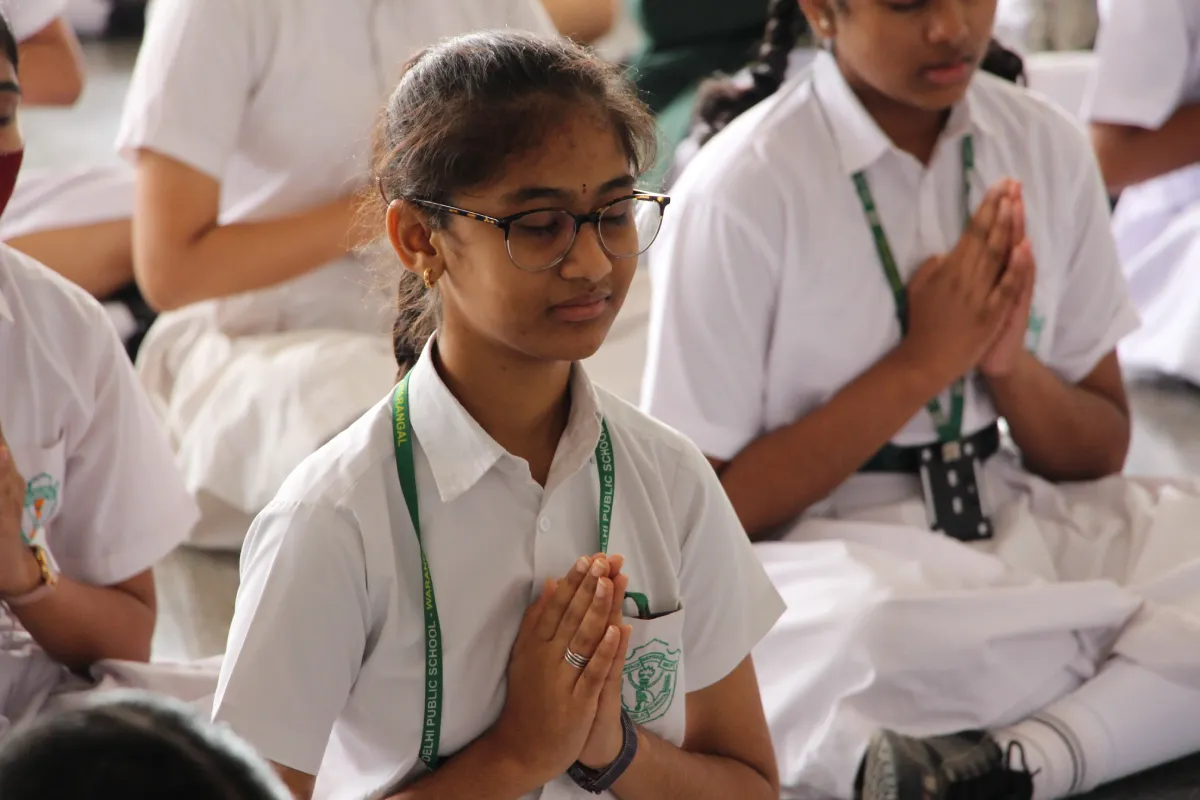 Students of DPS, Warangal in proper dress sitting on the ground and practicing breathing exercises with folded hands.