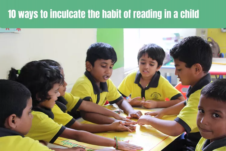 A group of children wearing yellow shirts are gathered around a table, engrossed in a shared activity. Three boys in yellow shirts sit on one side of the rectangular table, while on the opposite side, a larger group of children in matching shirts sits together. The scene is set against a plain background and features a banner with the text "10 ways to encourage the habit of reading in a child" hanging above the children. The focus shifts to one of the boys, who is smiling brightly at the camera with his hands resting on the table. Another boy in a yellow shirt also smiles at the camera from across the table.