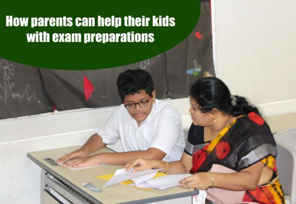 A young boy wearing glasses and a white shirt sits at a desk, preparing for his exam with his mother wearing a sari