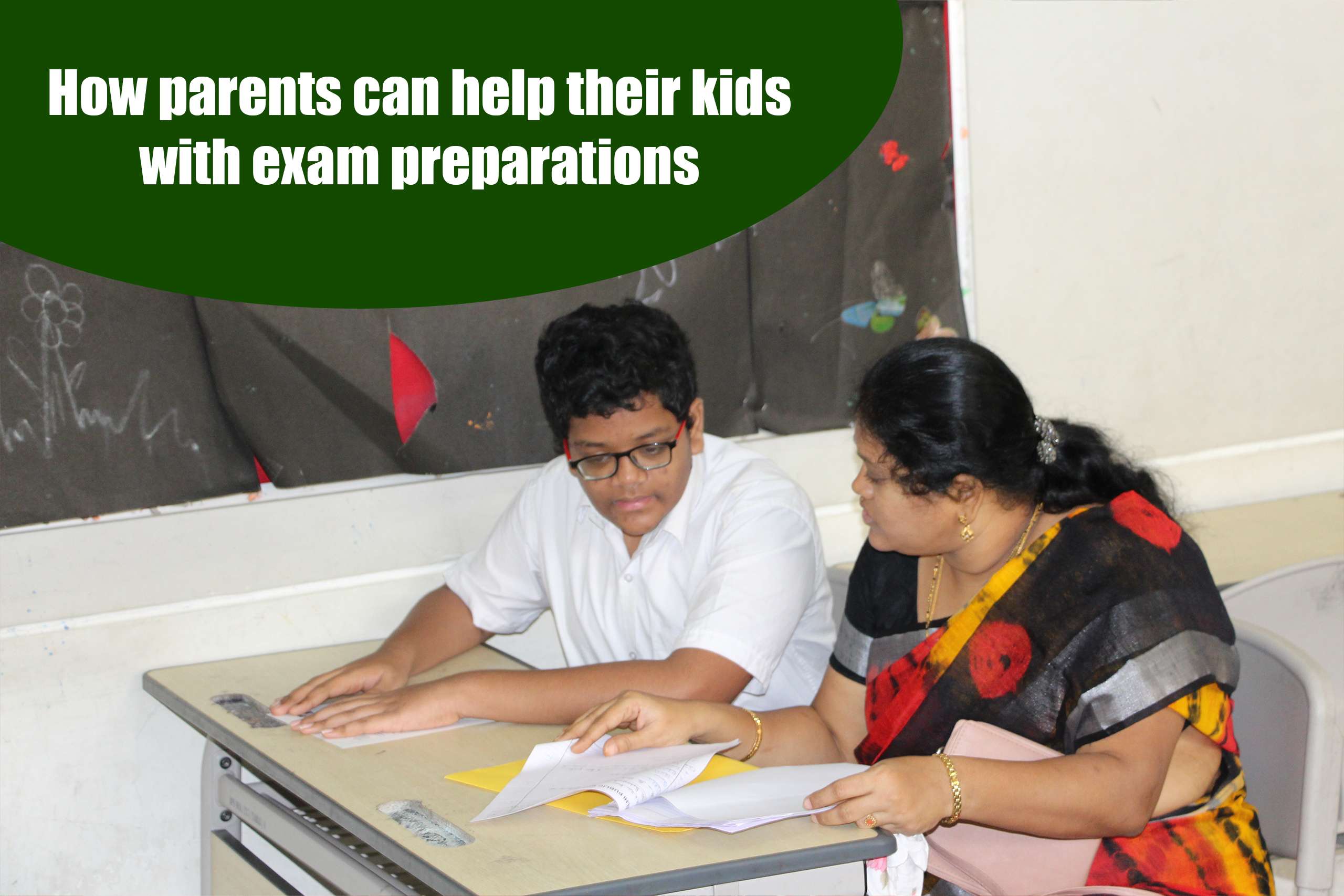 A young boy wearing glasses and a white shirt sits at a desk, preparing for his exam with his mother wearing a sari