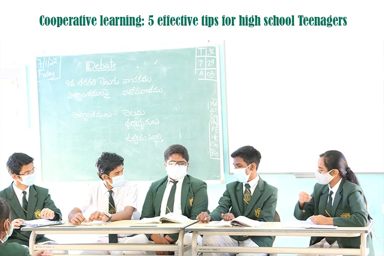 Five high school teenagers, wearing masks and school uniforms, are seated around a table in a classroom, engaged in a debate, demonstrating cooperative learning in action.
