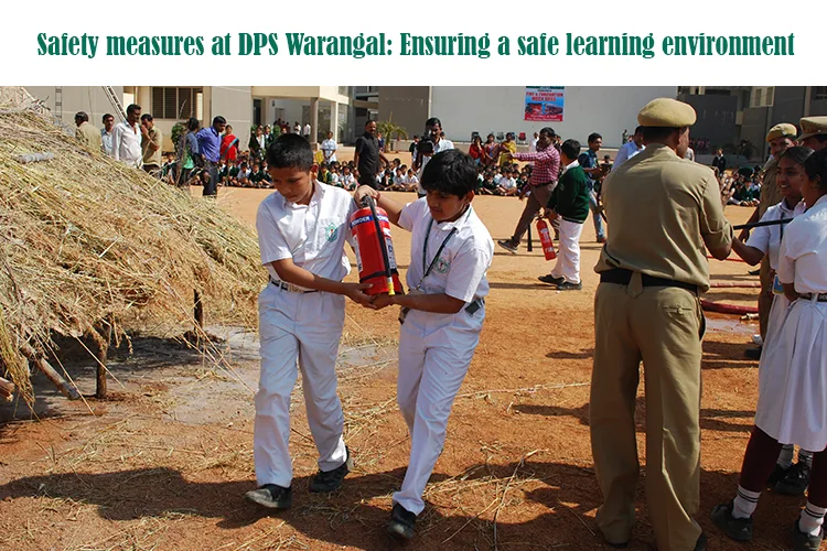 Two young male students in white uniforms are being instructed by an adult in a uniform on how to handle a fire extinguisher near a pile of straw, demonstrating safety measures at DPS Warangal