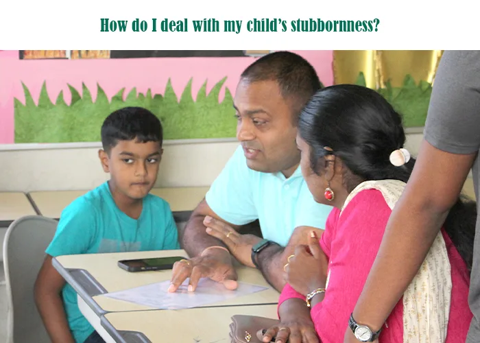 Parents talking to a teacher while a stubborn child sits beside them in a classroom, reflecting a discussion about managing a child’s stubborn behavior.